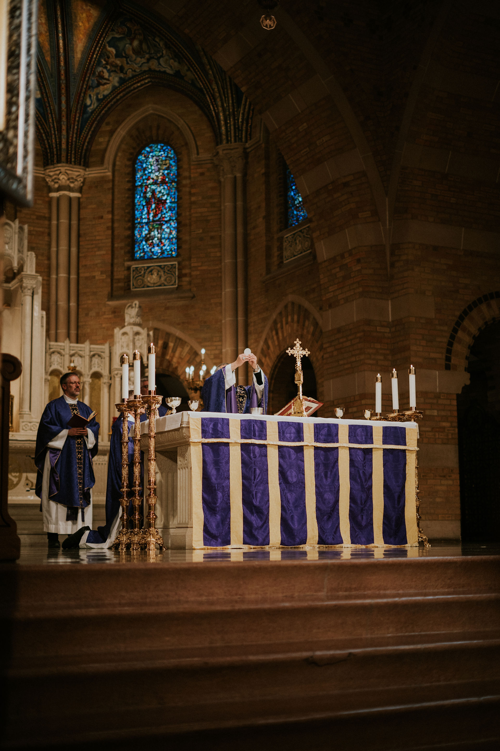 Priest in purple vestments raises a host at a church altar with lit candles. Stained glass windows and arches create a solemn atmosphere.