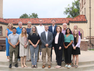 Twelve smiling adults pose on steps outside a brick building with a red tile roof, dressed in business casual.