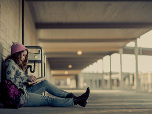 Girl in a pink beanie reads a book, sitting against a wall in a corridor. Her backpack rests beside her. The scene is calm and serene.