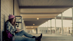 Girl in a pink beanie reads a book, sitting against a wall in a corridor. Her backpack rests beside her. The scene is calm and serene.