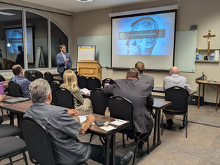 Man presenting to a classroom with a projection titled "The FBI's Behavioral Analysis Unit." Attendees seated, note-taking in a neutral setting.