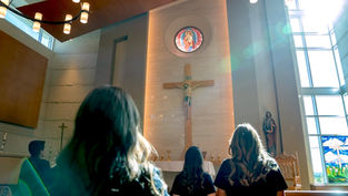 People in a church gaze at a cross under colorful stained glass, surrounded by soft light and wooden pews, creating a serene atmosphere.
