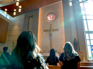 People in a church gaze at a cross under colorful stained glass, surrounded by soft light and wooden pews, creating a serene atmosphere.