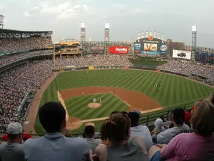 Fans watch a baseball game in a crowded stadium. The field is green with players in white. Large screens display ads. Clear sky above.