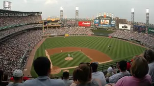 Fans watch a baseball game in a crowded stadium. The field is green with players in white. Large screens display ads. Clear sky above.