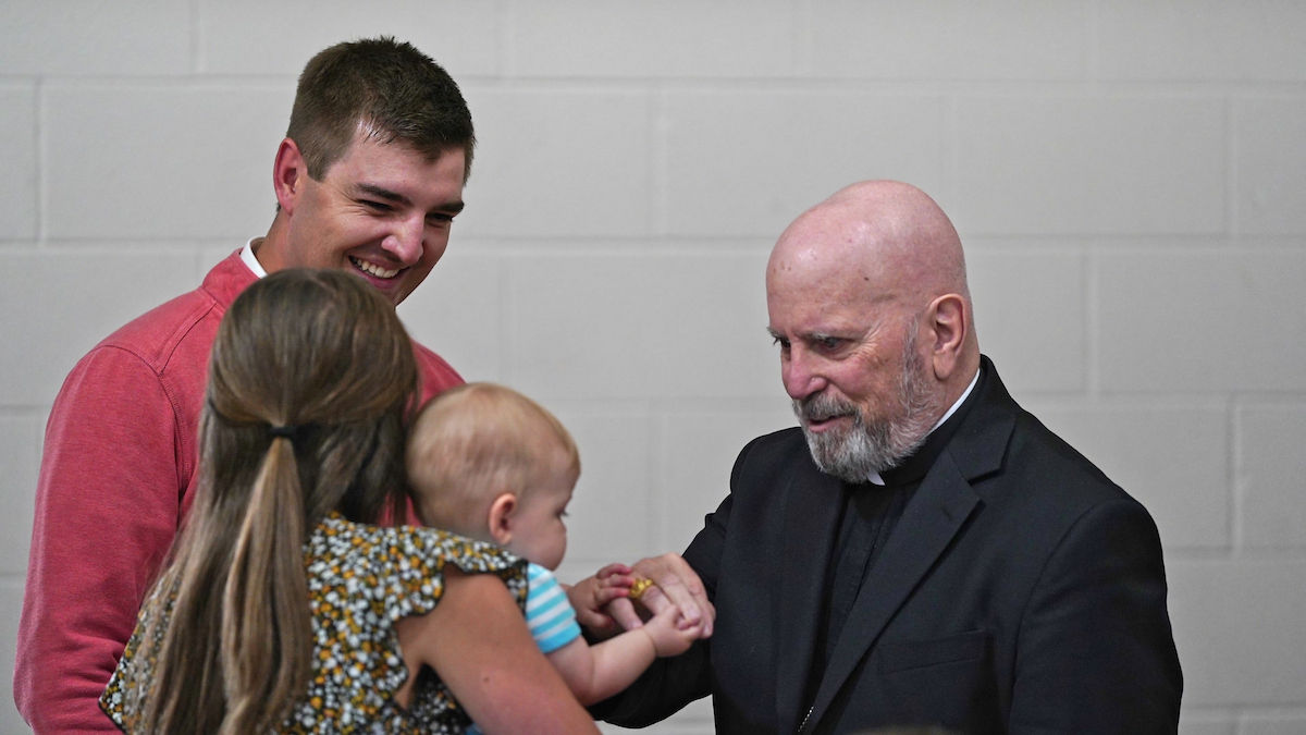 A man in a red sweater smiles as a woman holding a baby interacts with an elderly man in a black suit. Gray brick background.