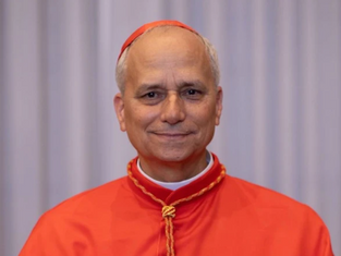 Smiling man in red religious attire with gold details, standing against a plain gray curtain background.