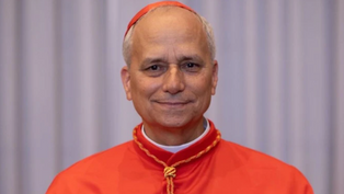 Smiling man in red religious attire with gold details, standing against a plain gray curtain background.