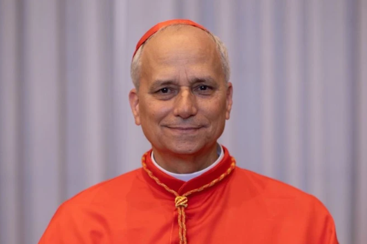Smiling man in red religious attire with gold details, standing against a plain gray curtain background.