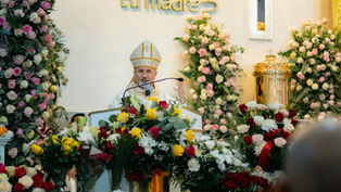 Bishop speaking at a podium surrounded by colorful flowers. Text "tu madre" on the wall. Church setting with ornate decor.