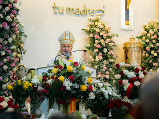 Bishop speaking at a podium surrounded by colorful flowers. Text "tu madre" on the wall. Church setting with ornate decor.