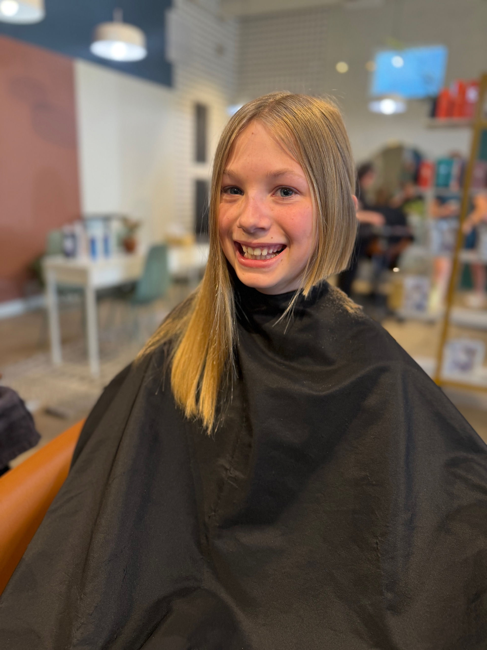 Smiling girl with freshly cut hair sits in a salon chair wearing a black cape.