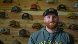A man with a beard wears a "Colorado" hoodie and cap, standing in front of a wooden wall displaying various hats. Mood: casual.