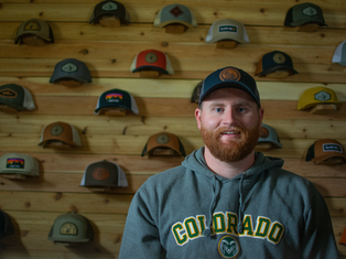 A man with a beard wears a "Colorado" hoodie and cap, standing in front of a wooden wall displaying various hats. Mood: casual.