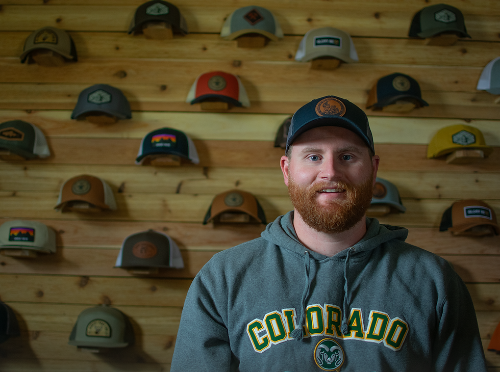 A man with a beard wears a "Colorado" hoodie and cap, standing in front of a wooden wall displaying various hats. Mood: casual.