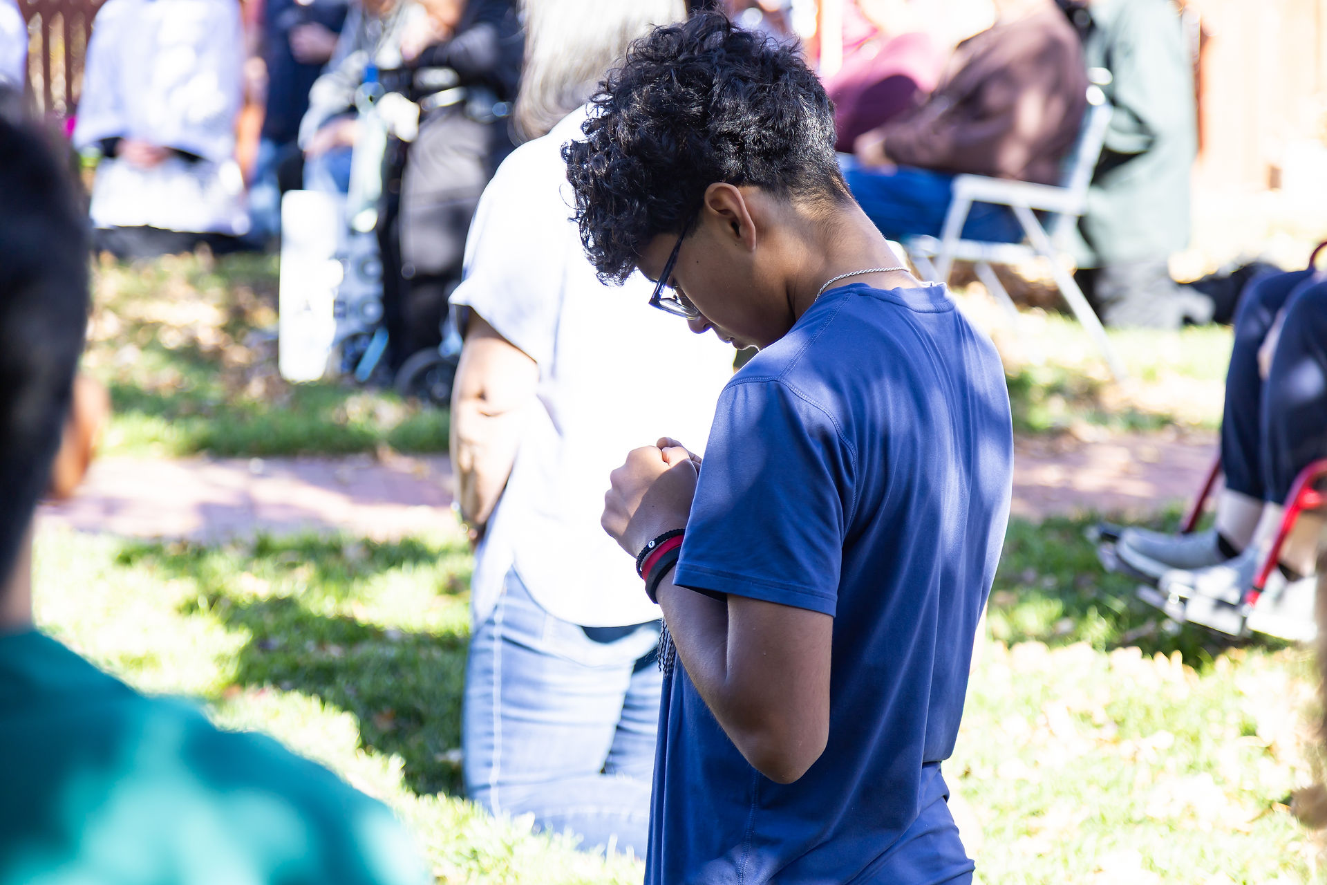Person in blue shirt looks down, standing in a grassy area with people sitting in the background. Sunny, outdoor gathering vibe.