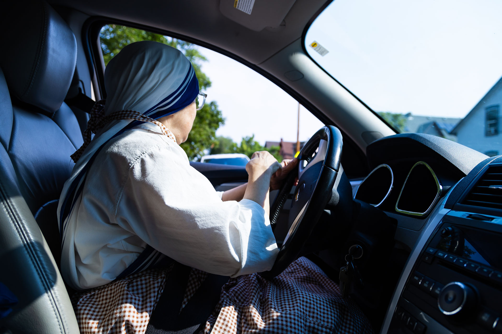 A person in a religious habit drives a car, with sunlight streaming through the window. Trees and houses are visible outside.