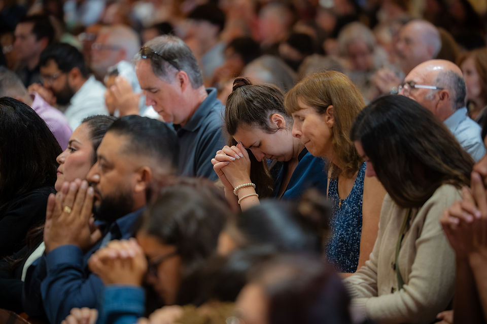 Crowd of diverse people sitting in an auditorium, eyes closed and hands clasped in prayer, solemn and reflective.