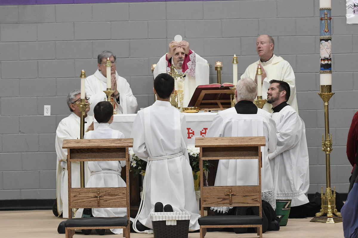 Clergy in white robes performing a religious ceremony with candles and an altar. One person holds up a wafer. Background has a brick wall.