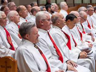 Rows of older men in white robes and red stoles sit solemnly in church pews during a religious ceremony.