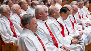 Rows of older men in white robes and red stoles sit solemnly in church pews during a religious ceremony.