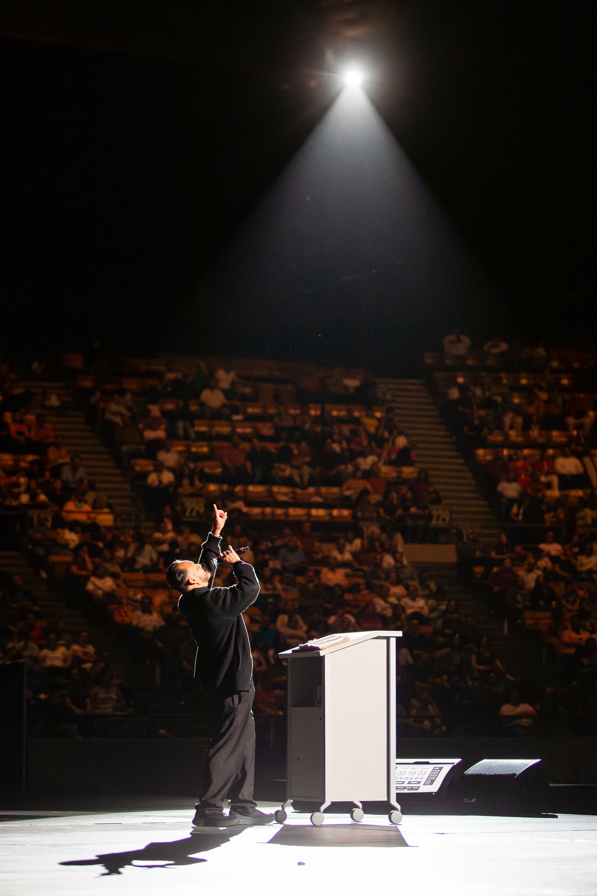 Speaker on stage, pointing upwards under a spotlight, addresses a large audience in a dimly lit auditorium. Energetic atmosphere.