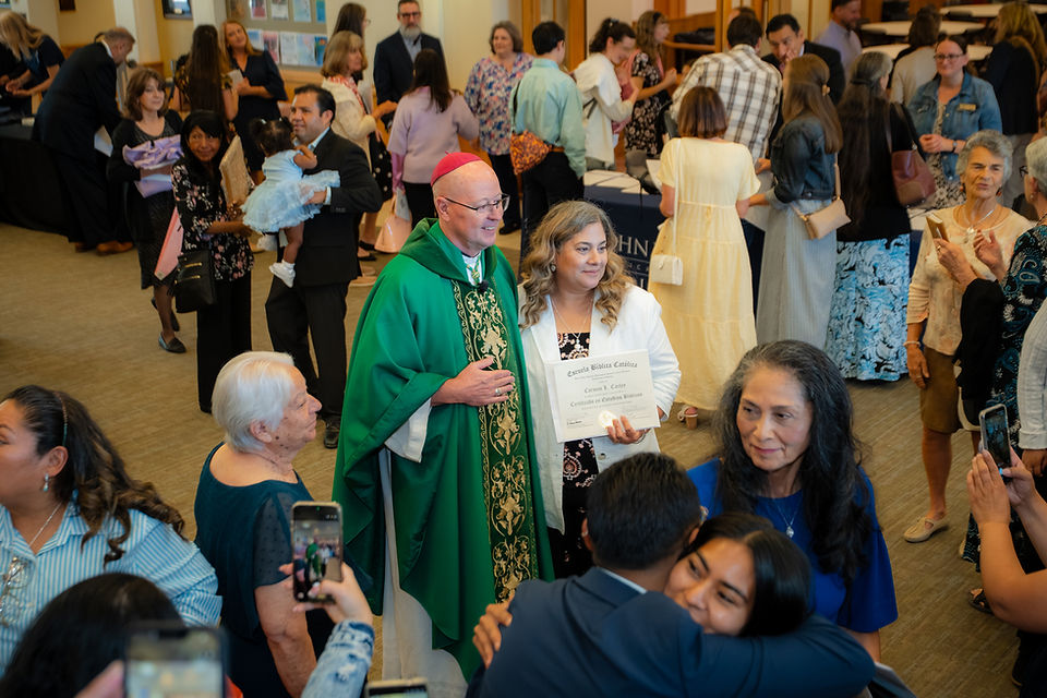 Bishop in green vestments poses with a woman holding a certificate at a crowded church celebration, with guests taking photos.