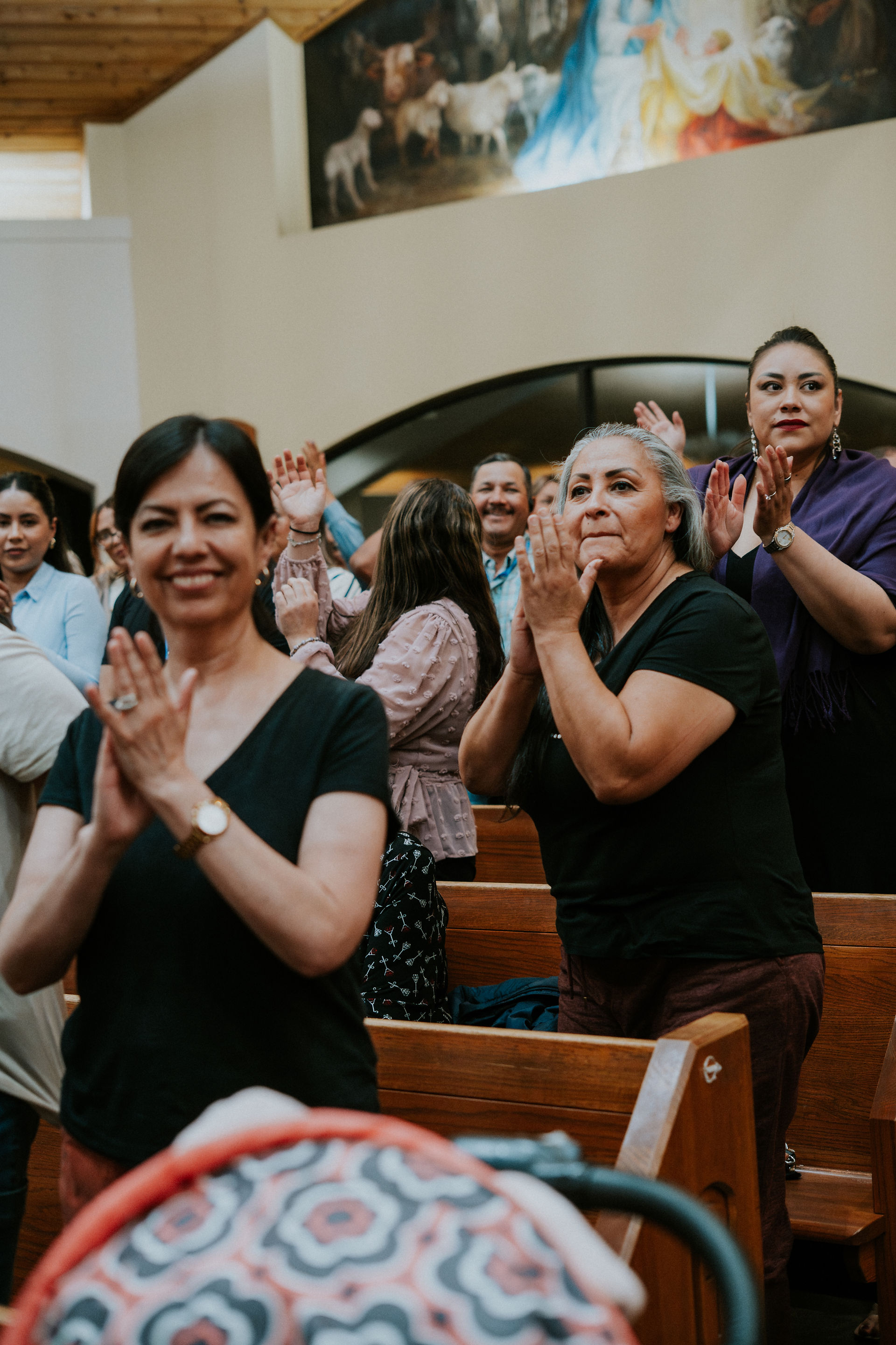 People clapping and smiling inside a church with wooden pews. A vibrant mural depicting animals is visible in the background.