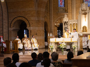 Priests perform a ceremony at a church altar, surrounded by clergy in ornate robes. Stained glass windows and candles add a solemn ambiance.