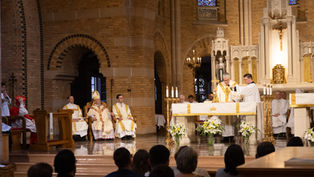 Priests perform a ceremony at a church altar, surrounded by clergy in ornate robes. Stained glass windows and candles add a solemn ambiance.