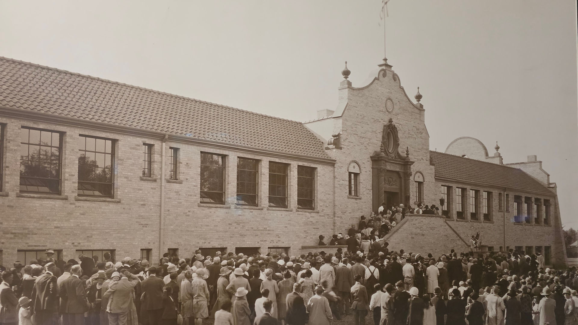 Sepia crowd gathered outside a brick building with ornate facade, lined windows, and steps at the entrance.