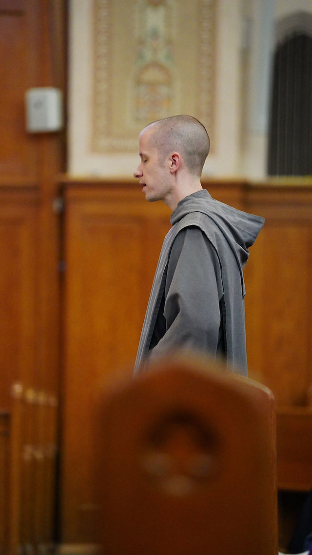 A person in a gray robe stands in a wooden church interior, with a calm expression. The background is ornate and warm-toned.