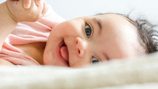 A smiling baby lies on a soft surface, wearing a pink shirt. The background is a light blur, adding a serene and joyful mood.