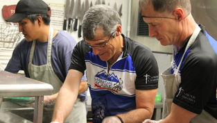 Three men wearing aprons work together in a kitchen. Two are in "Team Samaritan House" jerseys. Knives are visible on the wall behind them.