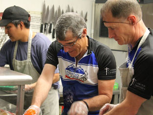 Three men wearing aprons work together in a kitchen. Two are in "Team Samaritan House" jerseys. Knives are visible on the wall behind them.
