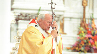 Pope St. John Paul II, vested in yellow and white robes, holds a cross, eyes closed in prayer. Background features blurred floral decorations. Peaceful atmosphere.