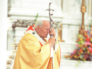 Pope St. John Paul II, vested in yellow and white robes, holds a cross, eyes closed in prayer. Background features blurred floral decorations. Peaceful atmosphere.