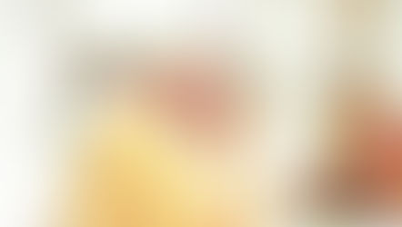 Pope St. John Paul II, vested in yellow and white robes, holds a cross, eyes closed in prayer. Background features blurred floral decorations. Peaceful atmosphere.
