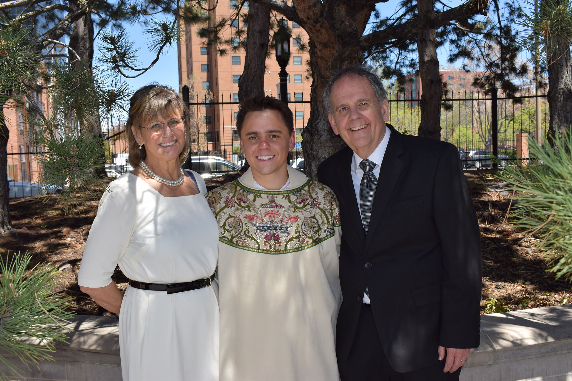 Three people smiling outdoors by trees, one in a floral robe, others in a dress and suit. Sunny day, buildings in the backdrop.