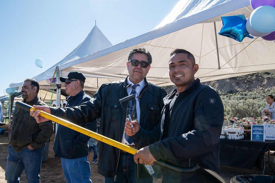 Two men smile while holding ceremonial shovels at an outdoor church groundbreaking event beneath white tents.