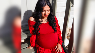 Pregnant woman in a flowing red dress sits on wooden steps, eyes closed, appearing serene. Sunlight casts soft shadows on a light wall.
