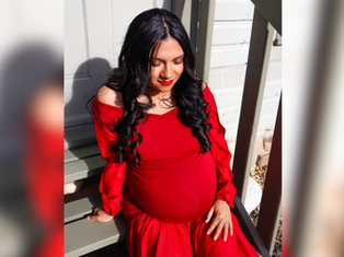 Pregnant woman in a flowing red dress sits on wooden steps, eyes closed, appearing serene. Sunlight casts soft shadows on a light wall.