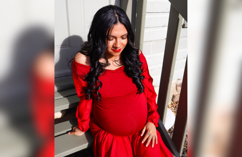 Pregnant woman in a flowing red dress sits on wooden steps, eyes closed, appearing serene. Sunlight casts soft shadows on a light wall.