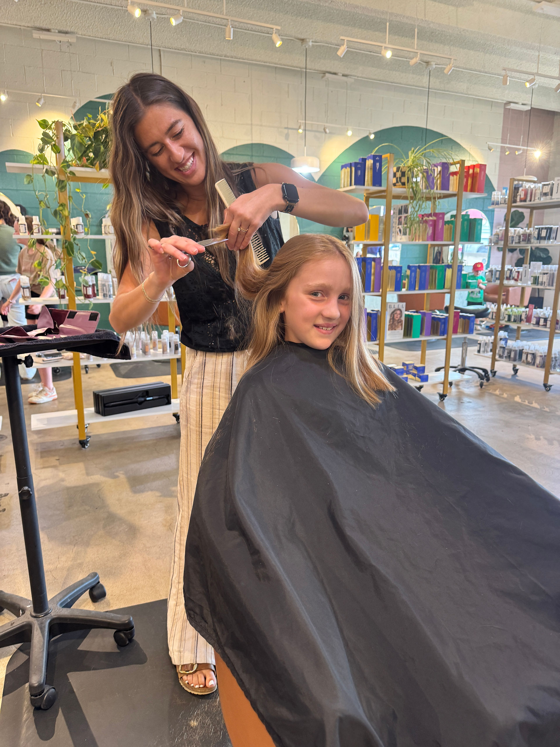 Hair stylist cuts a young girl’s long hair inside a bright salon with shelves of products in the background.