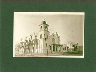 Sepia photo of a white church with bell towers and crosses on a quiet street, framed by leafless trees on a green background.