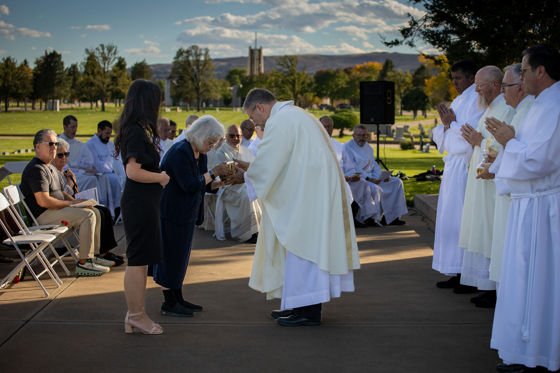 Clergy in white robes and attendees at an outdoor ceremony. An elderly woman gives a chalice to a priest. Scenic background with trees.