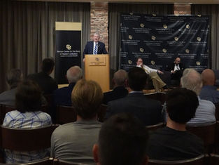 A speaker at a podium addresses an audience in a conference room. "Benson Center for the Study of Western Civilization" banner is visible.
