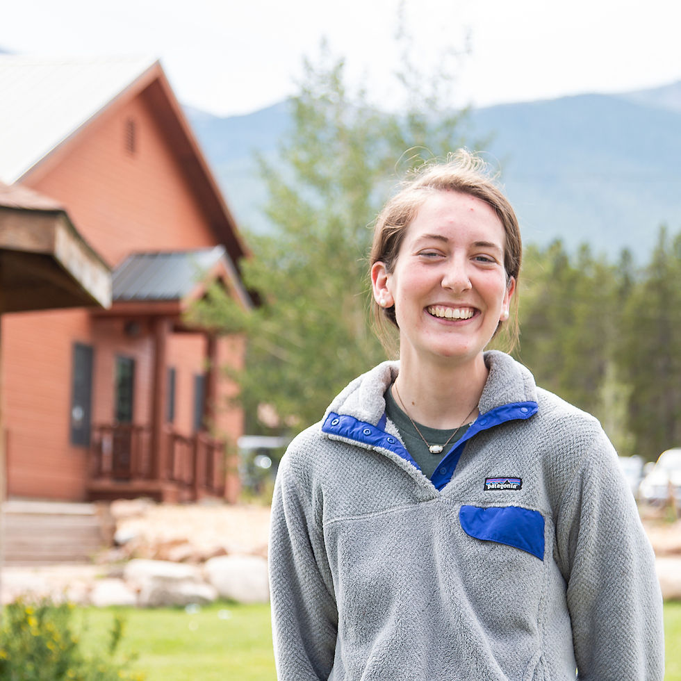 Smiling person in a gray fleece stands outdoors against a rustic cabin and mountain backdrop. Bright and cheerful mood.