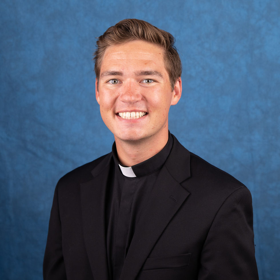 Smiling young man in a black clerical suit with a white collar, posed against a blue studio backdrop.