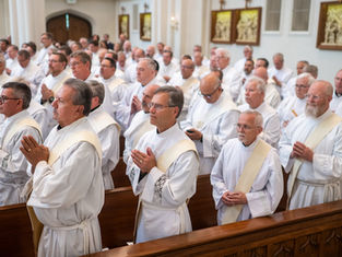 Clergy in white robes stand in a church, hands clasped in prayer. The setting is formal, with gold decor on the walls. Mood is reverent.